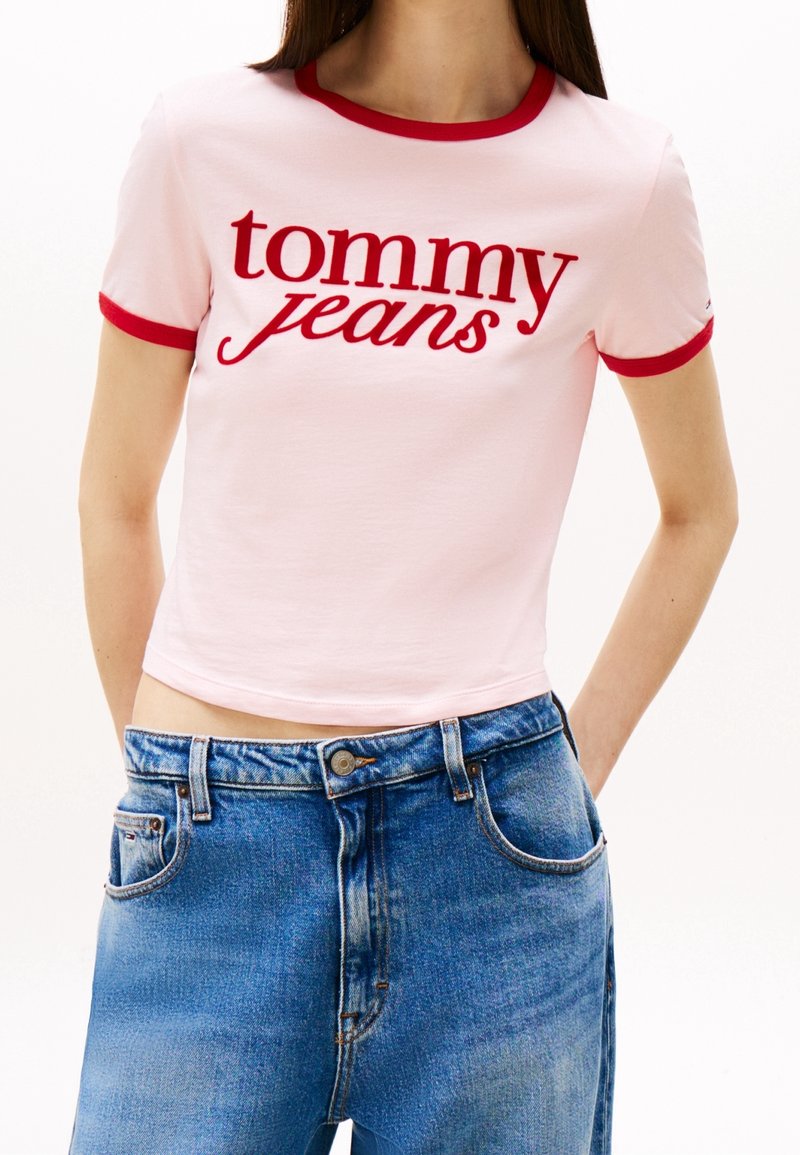 Pink cropped t-shirt with red contrast collar and sleeves, featuring “tommy jeans” in bold red text. Paired with high-waisted blue denim jeans.