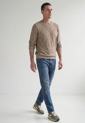 Mand går, iført beige sweater, blå jeans og grønne sneakers, mod en ensfarvet lys grå baggrund.