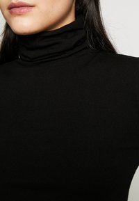 Kvinna med långt mörkt hår iförd en svart ribbad polotröja mot en ljus, enfärgad bakgrund.