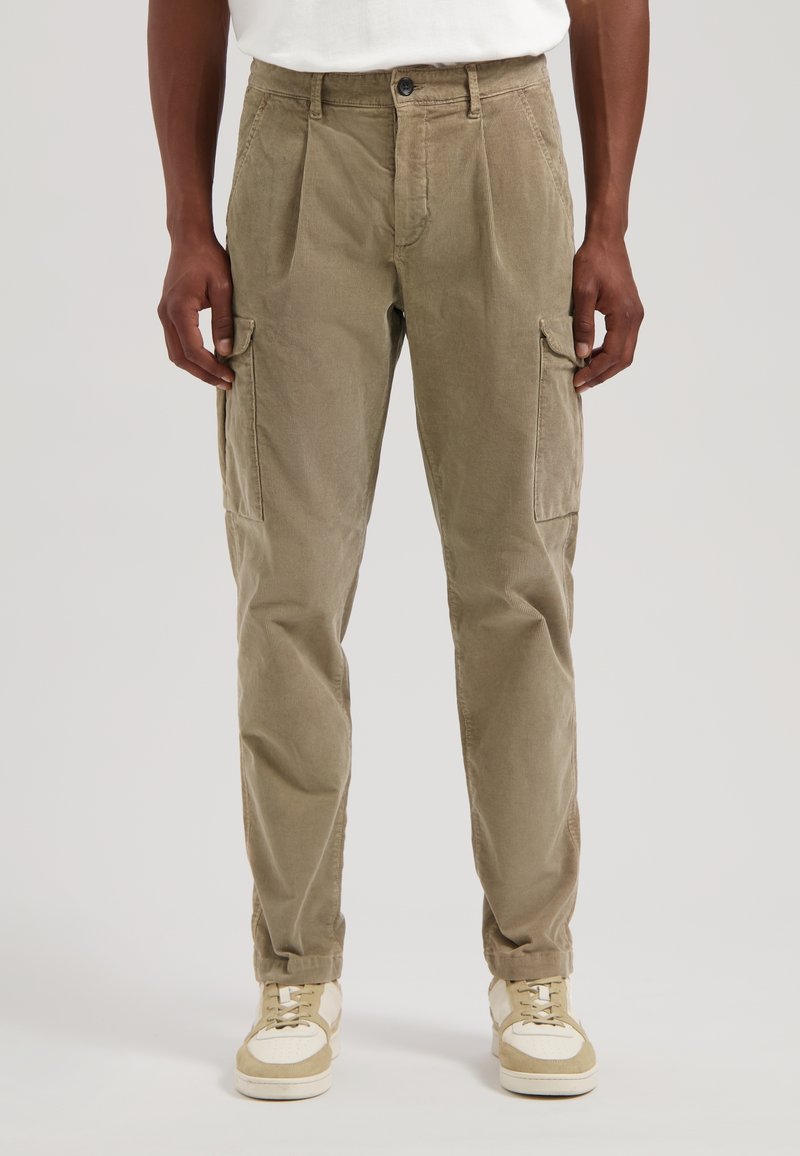 Khaki kordfløyel cargo bukser med folder, sidelommer og avsmalnende ben; kombinert med beige sneakers og en hvit t-skjorte.