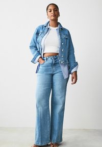 Lichtblauwe denimjack en bijpassende wijde jeans, met een cropped wit topje, oversized manchetten en een knopenlijst aan de voorkant.