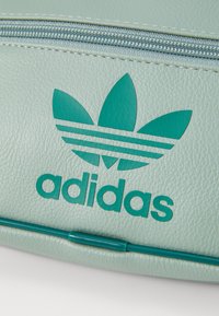 Světlozelená texturovaná kožená taška s tyrkysovým logem Adidas, která má tři pruhy ve tvaru listů a zipové uzávěr na vrchu.