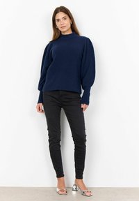 Pull en maille côtelée bleu marine avec un col montant et des manches bouffantes, associé à un jean noir ajusté et des sandales à talons argentées.