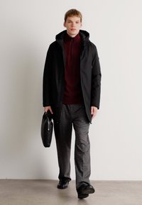 Jonge man die binnen loopt, gekleed in een zwarte hoodie, bordeauxkleurige trui, grijze broek, zwarte schoenen en een zwarte aktetas.