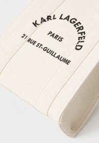 Beżowa torba z płótna z czarnym napisem "KARL LAGERFELD PARIS 21 RUE ST-GUILLAUME." Posiada wyszywane akcenty i wyraźny kształt.