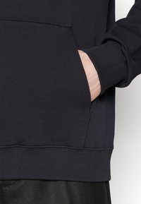 Hoodie noir avec une poche kangourou. Caractéristiques : poignets et ourlet côtelés. Texture lisse avec une coupe décontractée, montré de côté avec une main dans la poche.