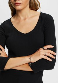 Frau mit verschränkten Armen, die ein schwarzes geripptes V-Ausschnitt-Top, ein silbernes Kettenarmband und mehrere silberne Ringe an ihren Fingern trägt.