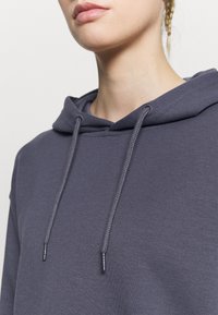 Sudadera con capucha gris de textura suave, con cordón y un corte relajado. Diseño liso sin logotipos ni patrones visibles.