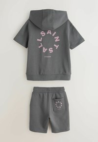 Sivá súprava s krátkym rukávom s kapucňou a kraťasmi, s ružovým kruhovým nápisom „SAINTS LONDON“ na chrbte a vrecku na kraťasoch.