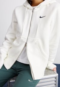 Vit zip-up hoodie i mjukt material med en svart Nike-logotyp, justerbara snoddar och framfickor, kombinerad med gröna byxor.