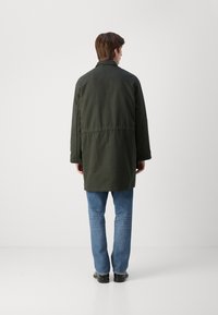 Lang, mørkegrøn frakke lavet af et glat, mat stof. Har en lige pasform, knaplukning og er kombineret med lyseblå jeans og sorte sko.