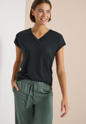 Femme souriante, portant un haut noir à manches courtes avec col en V et un pantalon vert à cordon, debout avec une main dans la poche sur un fond clair.