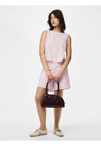 Jeune femme portant un haut en dentelle rose sans manches et un short, tenant un sac à main marron foncé, debout devant un fond blanc uni.