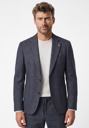 Man draagt een donkerblauw geruite colbert en bijpassende broek met een lichtgrijze trui met ronde hals eronder, staand tegen een witte achtergrond.
