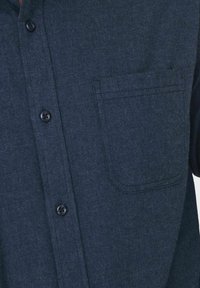 Chemise bleu foncé à boutons avec poche avant sur le côté gauche, montrant les détails des boutons et la texture du tissu.