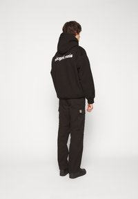 Schwarzer Hoodie mit dem Text "trahogami music" auf dem Rücken, kombiniert mit schwarzen Cargo-Hosen und schwarzen Schuhen. Das Outfit hat eine lässige Passform.