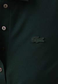 Polo shirt verde scuro realizzato in tessuto testurizzato, con colletto e tre bottoni argentati. Logo di coccodrillo verde ricamato sul petto.