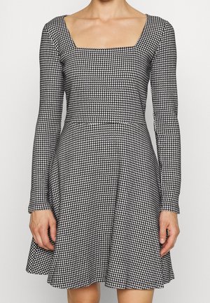 Žena v černobílých šatech s dlouhým rukávem, vzorem psí zub, čtvercovým výstřihem a rozšířenou sukní na jednoduchém pozadí.