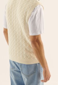 Krämfärgad kabelstickad ärmlös tröja med ribbade kanter, bärs över en vit t-shirt, i kombination med ljusblå jeans.