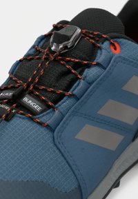 Plava atletska cipela s teksturiranim gornjim dijelom, elastičnim bungee vezicama, reflektirajućim trakama i crnim podstavljenim ovratnikom. Ima Gore-Tex brendiranje.