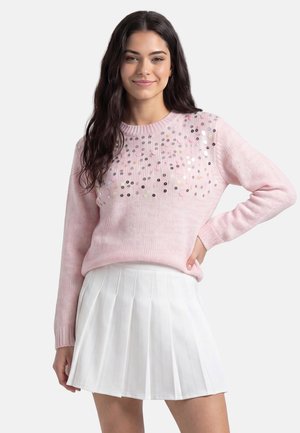 Donna con lunghi capelli scuri indossa un maglione rosa chiaro con paillettes e una mini gonna plissettata bianca, in piedi con una mano sui fianchi.