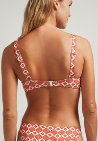 Top de bikini de rayas rojas y blancas con un diseño geométrico, tirantes ajustables y un cierre plateado en la parte posterior. Textura de tela suave.