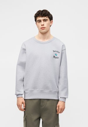 Fiatal férfi világosszürke pulóverben, melyen a „Service Works” felirat és egy kis tulipán grafika látható, egy egyszínű fehér háttér előtt áll.