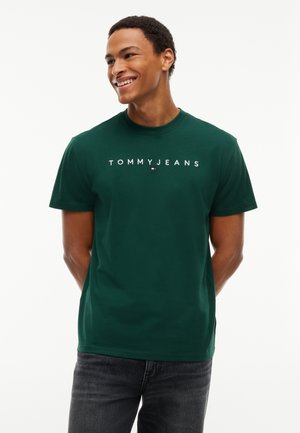 Junger Mann lächelt, trägt ein dunkelgrünes Tommy Jeans T-Shirt und dunkle Jeans, steht mit den Händen hinter dem Rücken vor einem weißen Hintergrund.