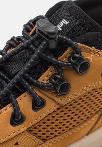 Tige en cuir marron avec des panneaux en mesh noir, dotée de lacets élastiques noirs et d'une fermeture à molette. Texturée avec un design respirant et une semelle en caoutchouc.