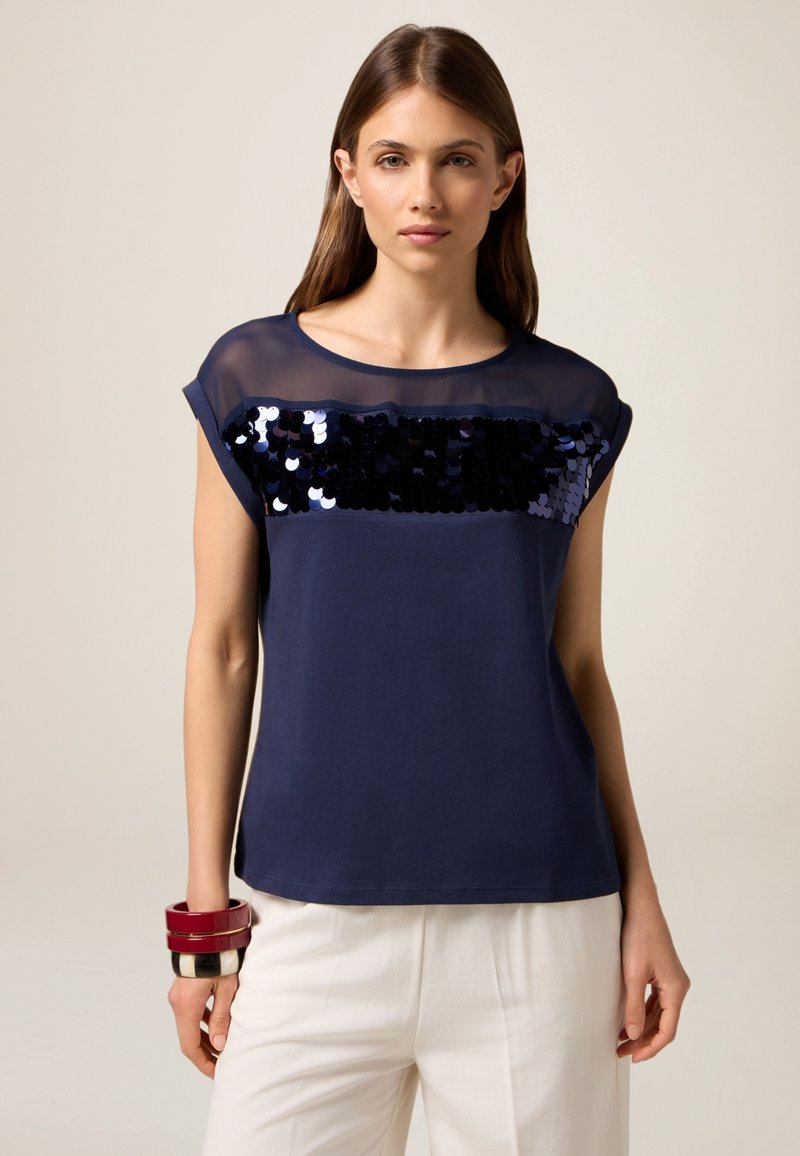 Top de manga corta azul marino con un panel transparente y un adorno de lentejuelas en el pecho. Tela suave con un corte relajado.