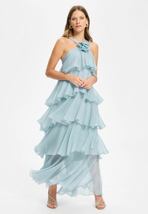 Femme portant une robe de soirée bleu clair à volants avec un col halter et des détails floraux, debout pieds nus sur fond blanc.