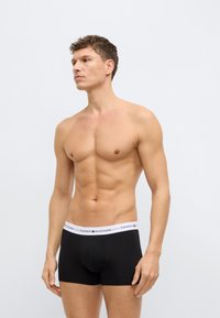 Black boxer shorts made of cotton, featuring a white elastic waistband with "TOMMY HILFIGER" logo, fitted design, and straight leg cut.