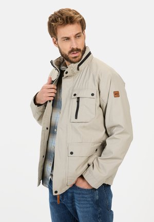Beige jacket with a high collar, multiple pockets, and black accents. Features a smooth texture and a zippered front. Worn over a plaid shirt.
