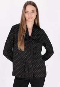 Bluză neagră cu buline albe, mâneci lungi, un fundiță legată la gât și o textură netedă a materialului. Design ajustat.