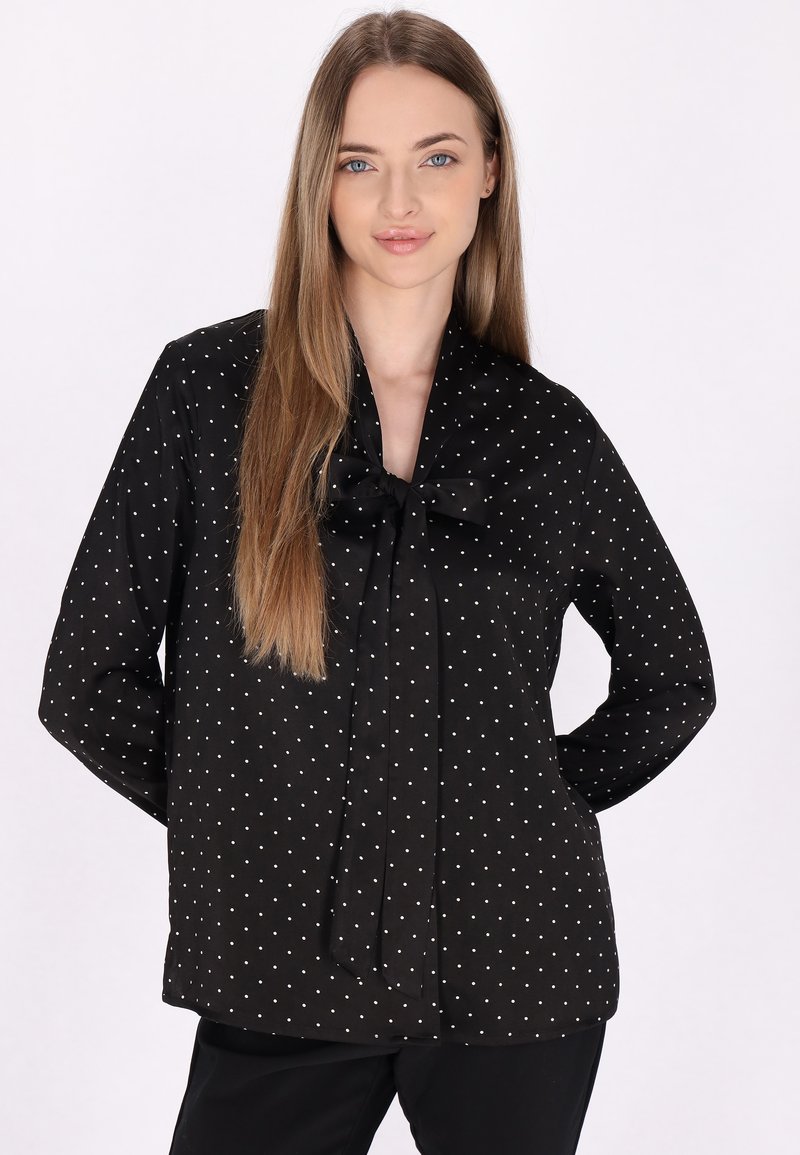 Bluză neagră cu buline albe, mâneci lungi, un fundiță legată la gât și o textură netedă a materialului. Design ajustat.