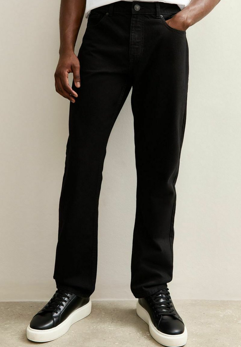 Vaqueros de mezclilla negra con corte recto, que cuentan con bolsillos frontales y cierre de botón. Combinados con zapatillas negras y acentos minimalistas en blanco.