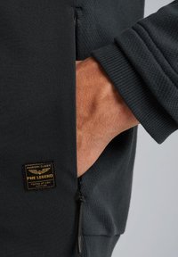 Zwarte ritssluiting jack van textuurstof, met een zichtbare pocket, rib gebreide manchetten en een PME Legend-logo patch aan de zijkant.