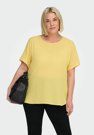 Vrouw met kort blond haar, gekleed in een geel T-shirt met korte mouwen en zwarte broek, die een zwarte handtas over haar schouder draagt.