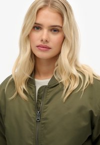 Oliwkowa kurtka bomberka z zamkiem błyskawicznym, dzianinowym kołnierzem i gładką fakturą. Nosi się ją na jasnym topie, który ma prosty design.