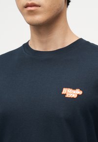 Άνδρας που φοράει σκούρο μπλε t-shirt με στρογγυλή λαιμόκοψη και πορτοκαλί και λευκό λογότυπο "JJ Studio 1990" στο αριστερό μέρος του στήθους.