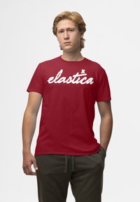 Rød kortærmet T-shirt med hvidt kursiv tekst "elastica", lavet af bomuld med en glat tekstur. T-shirten har en klassisk rund halsudskæring.