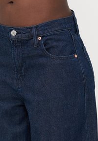 Jeans de mezclilla azul oscuro con un diseño clásico de cinco bolsillos, herrajes en tono cobre y un acabado de tela suave y texturizado.