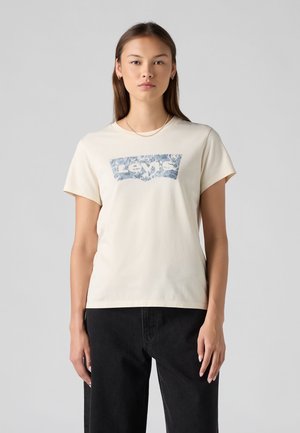 Młoda kobieta z prostymi włosami nosi kremowy t-shirt Levi's z niebieskim kwiatowym logo i czarne spodnie, stoi na jednolitym szarym tle.