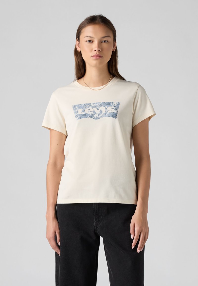 Νεαρή γυναίκα με ίσια μαλλιά φοράει κρεμ t-shirt Levi's με μπλε λουλουδάτο λογότυπο και μαύρο παντελόνι, στέκεται μπροστά σε απλό γκρι φόντο.
