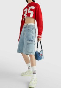 Červený zkrácený svetr s bílým síťovým detailem "35", modré džínové šortky s nášivkou "JEANS", bílé ponožky a tenisky s zelenými akcenty.