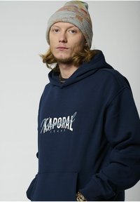 Sudadera azul marino de tejido suave, con un bolsillo frontal y un logo bordado en blanco. Combinada con un gorro de punto multicolor.