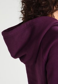 Sudadera morada con una textura suave, cuello ancho y una capucha adjunta visible en la imagen, con un diseño liso y sin costuras.