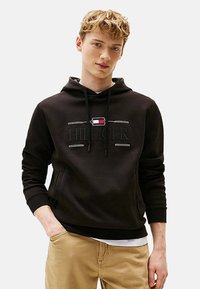 Sort bomulds-hoodie med snoretræk, der har "HILFIGER" logo, en lomme foran og ribbede manchetter. Bæres med khaki bukser. Simpelt design.