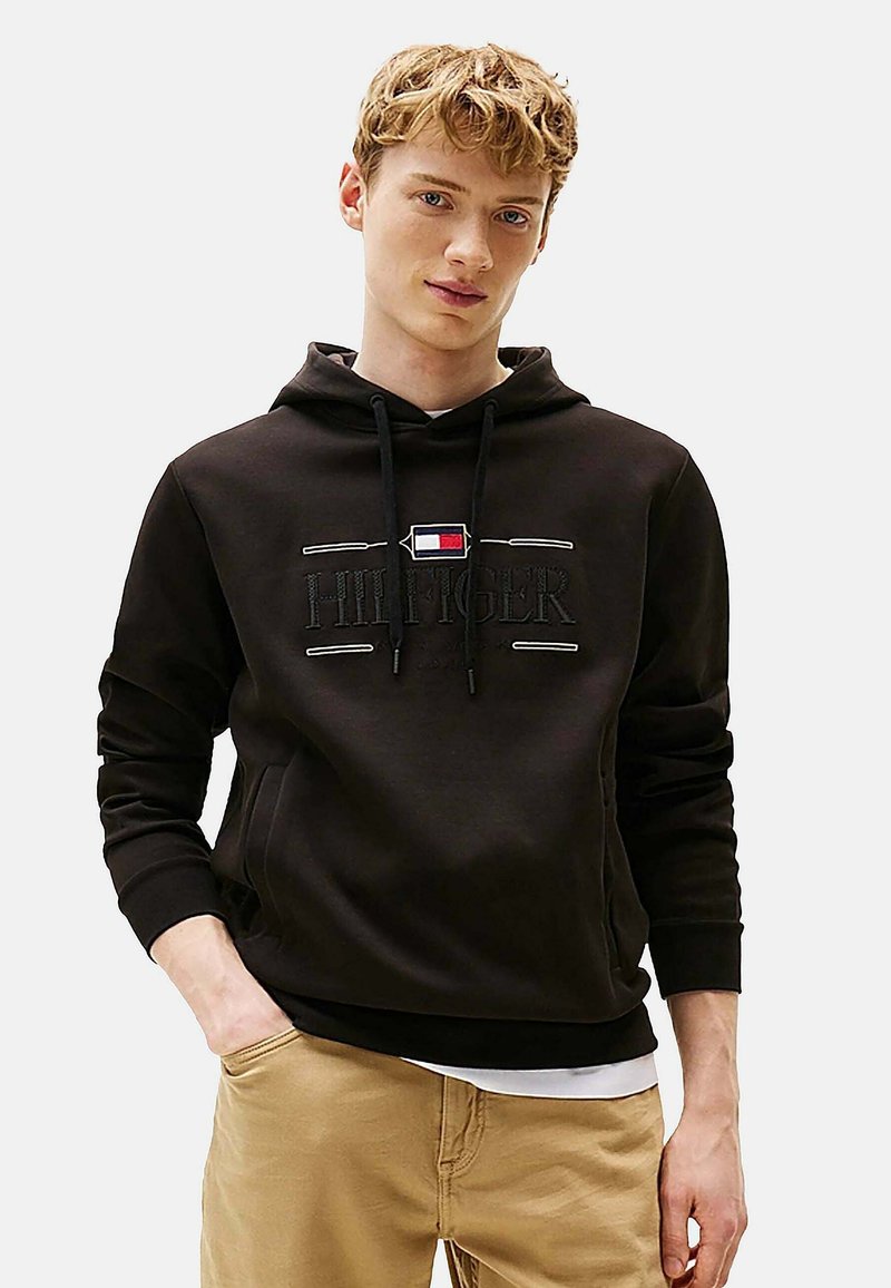 Sort bomulds-hoodie med snoretræk, der har "HILFIGER" logo, en lomme foran og ribbede manchetter. Bæres med khaki bukser. Simpelt design.