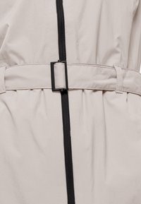 Lichtbeige jas met een zwarte ritssluiting en een riem met gesp-detail. Glad textuur, taille geaccentueerd, zichtbare stiksels langs de naden.